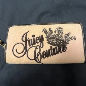  plånbok Juicy Couture med krona - Snygg beige plånbok från Juicy Couture med broderad logga och krona på framsidan. Utsidan är i mjuk mocka och detaljer i mörkbrunt skinn. Invändigt finns flera kortfack och har fina tryck på.  Den är lite använd så de finns några defekter i den som ex fläcken, skriv om du vill ha tydligare bilder!