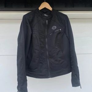 Svart Harley-Davidson jacka - Svart jacka från Harley-Davidson med coolt broderat logotyp på bröstet och stor 'Harley' text på ryggen. Jackan har dragkedjor framtill och på ärmarna.