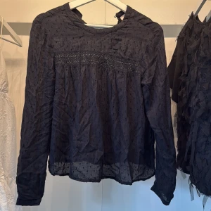 Svart blus med spets  - Säljer en svart blus med lång ärm och diskret prickigt mönster. Blusen har fina spetsdetaljer över bröstet och är tillverkad i ett tunt, lätt material som ger en luftig känsla. Perfekt för dig som gillar romantiska och stilrena plagg. Plagget är ostruket 
