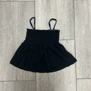 Svart smockad linnetopp Gina Tricot - Svart linnetopp från Gina Tricot med smockad överdel och tunna axelband. Toppen har en luftig, utställd nederdel och är tillverkad i 100% bomull. Perfekt för en chill och trendig look under varma dagar.