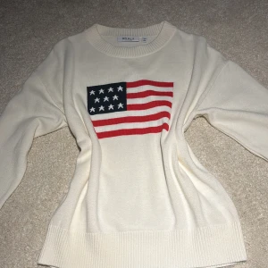 Vit stickad tröja med flagga från RYVLS - Vit stickad tröja från RYVLS med amerikansk flagga på bröstet. Tröjan har rund halsringning, ribbade muddar och är tillverkad i en mjuk blandning av bomull och akryl. Perfekt för dig som gillar klassisk och stilren design med en twist.