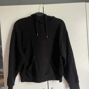 Svart hoodie från Lindex - En klassisk svart hoodie från Lindex med huva och snörning. Tröjan har en stor magficka och är tillverkad i mjuk bomullsblandning. Perfekt för chill dagar eller när du vill ha en enkel och stilren look.