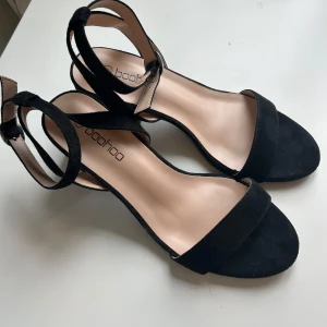 Svarta sandaler med klack från Boohoo - Använda 1 gång! Cirka 6 cm klack. Stilrena svarta sandaler från Boohoo med öppen tå och smal rem över foten. Skorna har en enkel klack och justerbar rem runt ankeln. Ytan är i mockaliknande material som ger en snygg och trendig look. Perfekta för varma dagar när du vill känna dig extra snygg.
