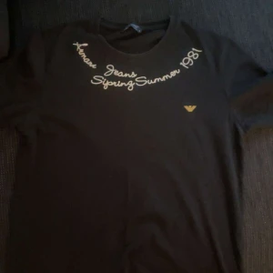 Svart t-shirt från Armani Jeans - Svart t-shirt från Armani Jeans med guldfärgad text och logga framtill. Snygg rund halsringning och tryck med 'Spring Summer 1981' samt exklusiv limited edition-detalj på ryggen. Perfekt för dig som gillar stilrena och ikoniska plagg.