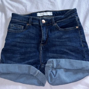 Mörkblå jeansshorts från Amisu - Snygga mörkblå jeansshorts från Amisu med klassisk femficksdesign och uppvikta benslut. Shortsen har guldiga sömmar, knapp och dragkedja framtill samt bälteshällor. Perfekta för varma dagar och enkel att matcha med din favorit-topp.
