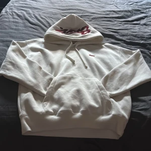 Vit hoodie från S.W.D. med röd brodyr - Säljer en clean vit hoodie från S.W.D. i storlek S. Tröjan har en stor röd broderad logga på huvan och en mindre logga på bröstet. Klassisk känguruficka och snörning i huvan. Mjuk bomullskvalitet som är skön att bära och passar perfekt till streetwear-stilen.
