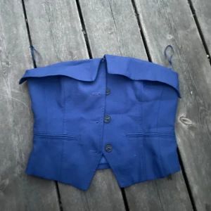 Blå korsett med knappar - Snygg blå korsett med knappar framtill och bred krage som ger en cool look. Axelbanden är tunna och toppen är croppad, perfekt för att sticka ut. Passar dig som gillar att mixa stilar och vill ha något unikt i garderoben.