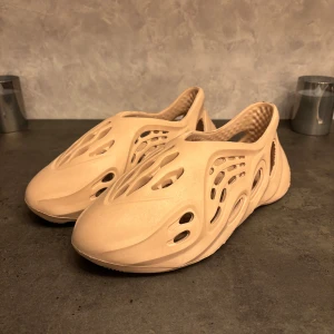 Beige adidas Yeezy Foam Runner - Säljer ett par beige adidas Yeezy Foam Runner med futuristisk design och många ventilerande hål. Skorna är tillverkade i syntetmaterial och har en platt sula med vågigt mönster undertill. Perfekt för dig som gillar unik stil och komfort.