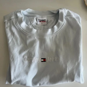 Ljusblå t-shirt Tommy Jeans - Fräsch ljusblå t-shirt från Tommy Jeans med broderad logga och text på bröstet. Klassisk rund hals och korta ärmar. Skön bomullskvalitet som känns mjuk mot huden. Perfekt för dig som gillar stilrena och enkla plagg med snygg branding.