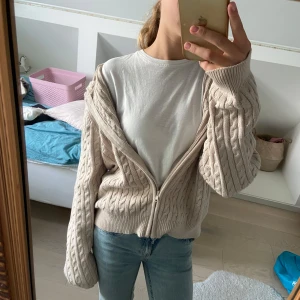Beige stickad cardigan jacka med dragkedja - Säljer en beige stickad cardigan jacka med dragkedja framtill. Jackan har ett snyggt kabelstickat mönster och ribbade muddar vid ärmslut och nederkant. Modellen är lite loose och har långärmade ärmar, perfekt för att slänga över en t-shirt. Från Lindex 