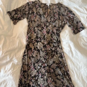 Svart blommig omlottklänning - Säljer en svart omlottklänning från rosebud med blommigt mönster i beige, rosa och vitt. Klänningen har v-ringning, korta ärmar och knälång passform. Materialet är mjukt och följsamt, perfekt för dig som gillar en feminin och bekväm stil.