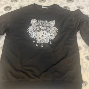 Kenzo tröja med silver tiger  - Svart sweatshirt från Kenzo med ikonisk silverbroderad tiger och logga på bröstet. Tröjan har rund hals, ribbade muddar och lång ärm. Perfekt för dig som vill ha en statement piece med cool Paris-vibe. Passar bättre som L