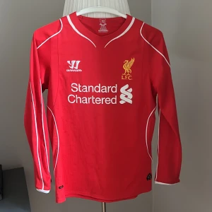Liverpool långärmad fotbollströja Warrior - Röd Liverpool FC fotbollströja från Warrior med lång ärm och vita detaljer längs sömmarna. Tryckt klubbmärke och sponsor på bröstet. Tillverkad i lätt och ventilerande polyester, perfekt för match eller träning. Klassisk V-ringad hals och snygg passform. 158 storlek 