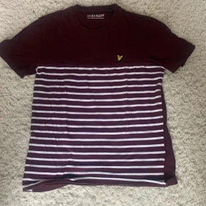 Vinröd randig t-shirt Lyle & Scott10-11 - Snygg vinröd t-shirt från Lyle & Scott med vita horisontella ränder på nedre delen och klassisk gul fågel-logga på bröstet. Rund hals och korta ärmar. Mjuk bomullskvalitet och normal passform, perfekt till jeans eller shorts.10-11 år