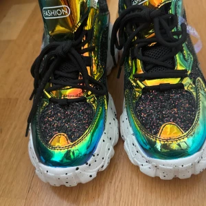 Glittriga chunky sneakers från SHEIN - Säljer ett par riktigt coola chunky sneakers från SHEIN med glittriga svarta paneler och metallic regnbågsfärgade detaljer. Skorna har tjock vit sula med svarta prickar, svarta snören och en unik futuristisk vibe. Perfekt för dig som vill sticka ut med din stil.