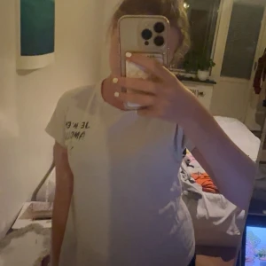 Vit t-shirt med tryck Je m'en Amour - Säljer en vit t-shirt med svart tryck på bröstet där det står 'Je m'en Amour' och en liten stjärna under texten. T-shirten har rund hals och korta ärmar, klassisk passform och är gjord i mjukt bomullstyg. Perfekt för dig som gillar enkla men snygga statement-plagg. passar xs