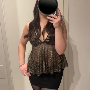 Glittrig svart och guld linnetopp - Säljer en glittrig linnetopp i svart och guld med tunna axelband och djup v-ringning. Toppen har ett flowigt fall och är dekorerad med paljetter som ger en riktigt festlig vibe. Knytning i ryggen för justerbar passform. Perfekt för att sticka ut på kvällen!