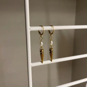 Guldiga örhängen med pärldetalj -  I rostfritt stål och handgjort. Snygga örhängen i guld med hängande konformad detalj och en liten vit pärla ovanför. Perfekt för dig som gillar att sticka ut med coola accessoarer. De har en modern och edgy vibe som passar till många olika stilar.