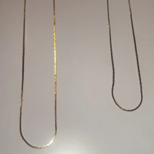 Två tunna guldiga halsband - Säljer två stilrena och tunna halsband i guldton. Kedjorna har en platt och glansig yta som reflekterar ljuset snyggt. Perfekt för dig som gillar minimalistiska smycken och vill ha något som funkar till alla outfits. Båda har klassisk låsning.