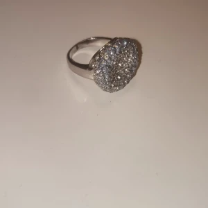 Glittrig silverring med stenar - Säljer en rund, glittrig silverring med massor av små gnistrande stenar på ovansidan. Ringen har en bred och blank ringbas i silverfärg och en stor, upphöjd dekoration som verkligen drar blickarna till sig. Perfekt för dig som gillar att sticka ut med dina accessoarer.