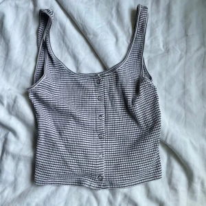 Vit och svart rutig croppad linnetopp - Säljer en croppad linnetopp i vit och svart med rutigt mönster. Toppen har knappar framtill och breda axelband, perfekt för dig som gillar en chill och trendig stil. Materialet känns mjukt och lätt mot huden.