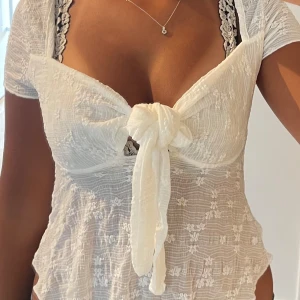 Vit blus med knyt och spetsdetaljer - Supersöt vit blus med knyt framtill och transparent spetsmönster. Blusen har korta ärmar och v-ringad hals, med romantiska detaljer och en feminin look. Perfekt för dig som gillar att sticka ut med snygga detaljer och lätt material.