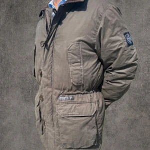 Brun parkajacka från Morris - Parkajacka från Morris i mörkbrunt med klassisk design och stora fickor framtill. Jackan har emblem på ärmen och snygga detaljer som knappar och dragkedja. Perfekt för kyliga dagar med sin vadderade insida och robusta känsla.