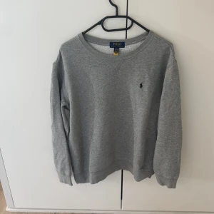 Grå sweatshirt från Polo Ralph Lauren - Snygg grå sweatshirt från Polo Ralph Lauren med klassisk rund hals och broderad marinblå logga på bröstet. Tröjan har ribbade muddar vid ärmslut och nederkant, och är tillverkad i mjuk bomullsmix som är perfekt för chill dagar.