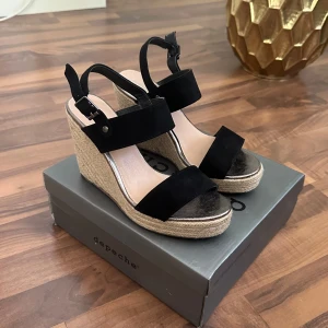 Svarta kilklack-sandaler från Depeche - Snygga svarta sandaler med kilklack från Depeche. Ovandel i mocka med breda remmar och justerbart spänne runt ankeln. Klacken är täckt med flätad jute för en chill sommarkänsla. Platt sula framtill och öppen tå ger en fräsch look till dina outfits.