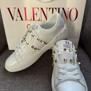 Valentino Rockstud Untitled sneakers - Snygga vita Valentino Rockstud Untitled sneakers med guldiga nitar och klassisk snörning. Skorna har en rund tå och platt sula, tillverkade i lyxigt skinn. Perfekt för dig som vill sticka ut med exklusiva detaljer och stilren design.