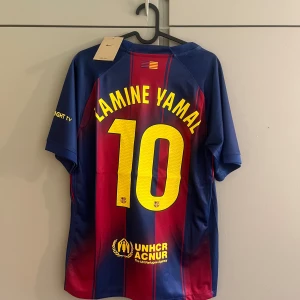 FC Barcelona Lamine Yamal tröja Nike - Säljer en officiell FC Barcelona matchtröja med Lamine Yamal och nummer 10 på ryggen. Tröjan är kortärmad, har klassiska blå och röda vertikala ränder, gula detaljer och klubbmärke på bröstet. Materialet är lätt och andas, perfekt för fotboll eller häng med kompisar.