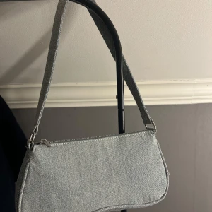 Jeans axelväska från H&M  - Säljer en stilren ljusblå axelväska i jeansmaterial. Väskan har en enkel, avlång form med dragkedja upptill och matchande axelrem. Sparsamt använd, som ny! 
