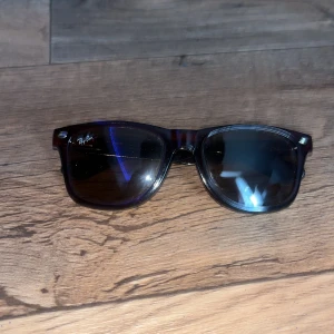 Bruna Ray-Ban Wayfarer solglasögon - Snygga bruna Wayfarer solglasögon från Ray-Ban med klassisk rektangulär form och mörka glas. Ramen är i blank plast och har diskret Ray-Ban-logga på linsen. Perfekt för dig som vill ha en tidlös och cool look. Andvänds max 10 gånger.