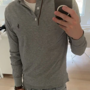Grå långärmad pikétröja från Polo Ralph Lauren - Säljer en stilren grå långärmad pikétröja från Polo Ralph Lauren med klassisk krage och knappar framtill. Tröjan har den ikoniska Polo-loggan broderad på bröstet och är gjord i mjuk bomull. Perfekt för dig som gillar en clean och enkel look.