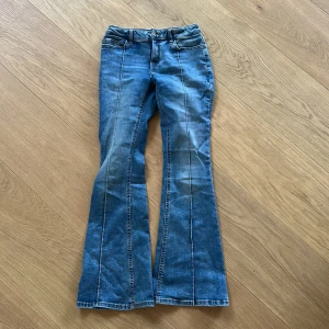 Blå bootcut jeans från Lindex, strl 152 - Snygga blå jeans från Lindex i storlek 152 med bootcut-ben och klassiska fickor både fram och bak. Jeansen har ljus tvätt och markerade sömmar längs benen för en cool look. Perfekta för dig som gillar retrovibbar och bekväma jeans.