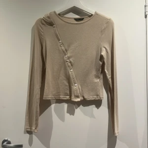 Långärmad topp med knappar - Säljer en beige långärmad topp från SHEIN i storlek M. Toppen har en cool asymmetrisk knappdetalj framtill och är croppad i modellen. Materialet känns mjukt och lätt. 