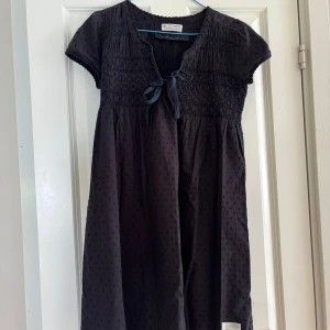 Svart klänning Odd Molly med knyt - Supersöt svart kortklänning från Odd Molly med prickigt mönster och smock upptill. Klänningen har korta ärmar, v-ringning och ett fint knytband framtill. Materialet är mjuk bomull och nederkanten pryds av en Odd Molly-label. Perfekt för dig som gillar detaljer och skön passform.