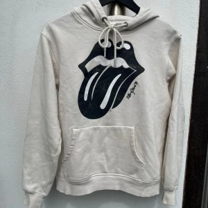 Rolling Stones hoodie H&M - Stilren beige Rolling Stones hoodie från H&M i storlek XS med ikoniskt Rolling Stones-tryck på bröstet och stor logga på ryggen. Tröjan har huva med snörning, känguruficka. Är tillverkad i bomull och polyester. Perfekt för dig som gillar bandmerch och streetstyle. Använd men väl bevarad o i fint skick, hel o ren.
