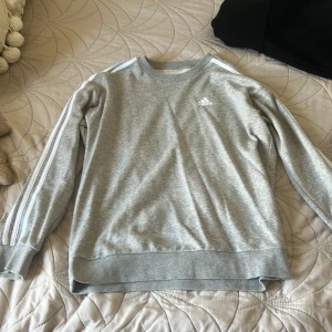 Grå Adidas sweatshirt - Säljer en grå sweatshirt från Adidas med klassiska vita ränder längs ärmarna och broderad logga på bröstet. Tröjan har rund hals, ribbade muddar och är långärmad. 