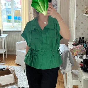 Grön skjorta med snörning och fickor - Snygg grön skjorta med korta ärmar, snörning i midjan som du kan dra åt utifrån preferens. Så fin färg och snygg till jeans, dock aldrig kommit till användning. Storlek XL, men somsagt väldigt justerbar. 