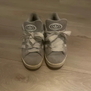 Grå Adidas Campus sneakers strl 38 - Säljer ett par grå Adidas Campus sneakers med klassiska vita ränder på sidan och Adidas-logga på plösen. Skorna har rund tå, platt sula och breda vita snören. Ovandelen är i mjuk mocka som ger en chill vibe. Perfekt för dig som gillar retrostil och streetwear. Kom med förslag på pris 