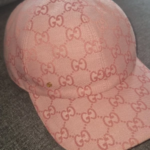 Rosa keps med GG-mönster - Snygg rosa keps med ikoniskt GG-mönster över hela kepsen. Justerbar rem bak i rosa skinn med guldfärgad metallring. Kepsen är tillverkad i bomull och har en klassisk böjd skärm. Perfekt för dig som vill sticka ut med lyxig streetstyle.