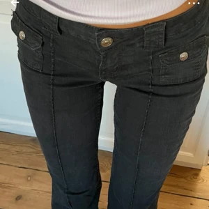 Svarta bootcut jeans med fickdetaljer - Säljer ett par svarta jeans med bootcut passform och snygga fickor med lock och silvriga knappar framtill. Byxorna har låg midja och markerade sömmar längs benen. Materialet är klassiskt jeans och de har dragkedja och knappstängning.