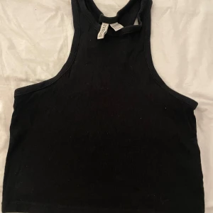 Svart ribbad linnetopp från H&M Divided - Svart croppad linnetopp från H&M Divided med ribbad struktur. Toppen har en hög halsringning och är ärmlös med snygg racerback-design. Perfekt att styla med jeans eller kjol för en chill och trendig look.