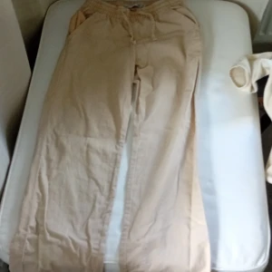 Beige linnebyxor från 157, strl 140 - Snygga beige linnebyxor från 157 med elastisk midja och snörning. Byxorna har en avslappnad passform och bakficka, perfekta för chill dagar. Materialet är linne som ger en skön och luftig känsla. Passar dig som gillar enkel och clean stil.