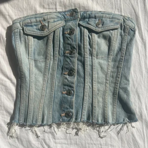 Ljusblå jeanskorsett med knappar - Säljer en ljusblå korsett i jeansmaterial med snygga metallknappar framtill och två dekorativa fickor. Toppen har fransig nederkant och smockad rygg för extra komfort. Perfekt för dig som vill ha en edgy och cool look.