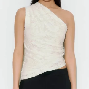 Vit spetsig oneshoulder topp - Säljer en vit topp med spetsdetaljer och oneshoulder-design. Toppens asymmetriska form ger en cool och modern vibe, och spetsen gör den extra feminin. Perfekt att styla med svarta jeans eller kjol för en trendig look.
