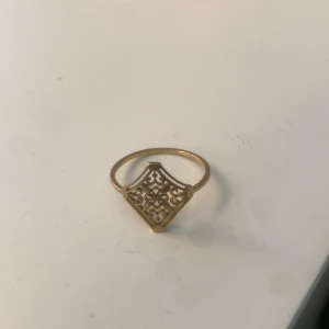 Guldfärgad ring med blommigt mönster - Snygg guldfärgad ring med ett unikt blommigt mönster i en fyrkantig form. Ringen har tunna sidor och ett detaljerat utskuret motiv som ger en cool och personlig vibe. Perfekt för dig som gillar smycken med lite extra karaktär.