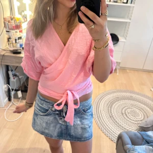 Rosa omlottblus med knyt i midjan - Säljer en ljusrosa omlottblus med knyt i midjan. Blusen har en v-ringad design och trekvartsärmar med lite puffig känsla. Materialet är mjukt och luftigt, perfekt för varmare dagar. Snygg att matcha med jeans eller kjol för en fräsch look.