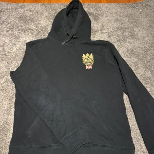 Svart hoodie från Morris med tryck - Svart hoodie från Morris med snyggt guldfärgat emblem och brittisk flagga tryckt på bröstet. Tröjan har huva med snörning, känguruficka och ribbade muddar. Perfekt för dig som gillar klassisk stil med en twist.